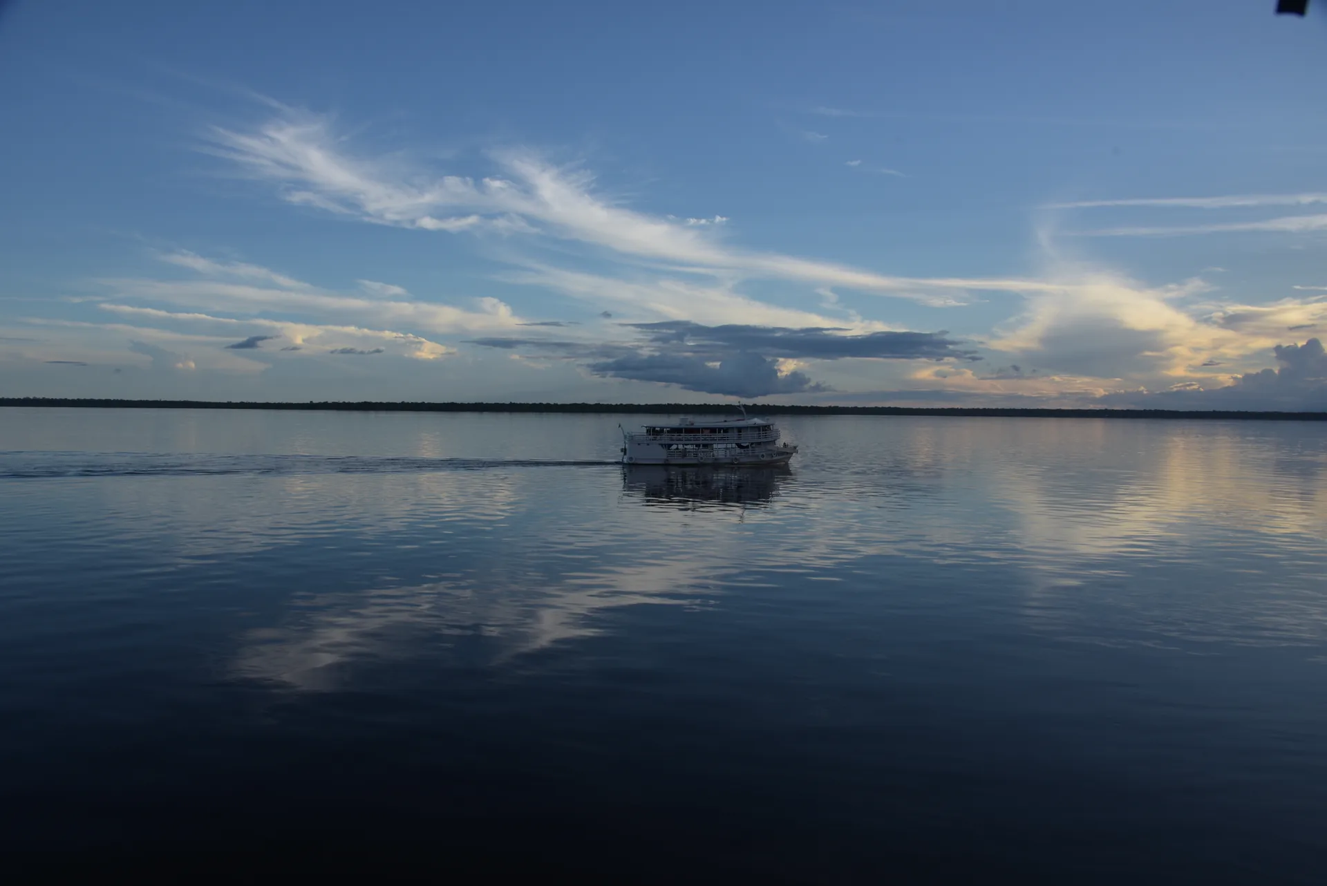 Barco regional navegando num rio largo e calmo que reflete o céu com nuvens finas, na Amazônia
