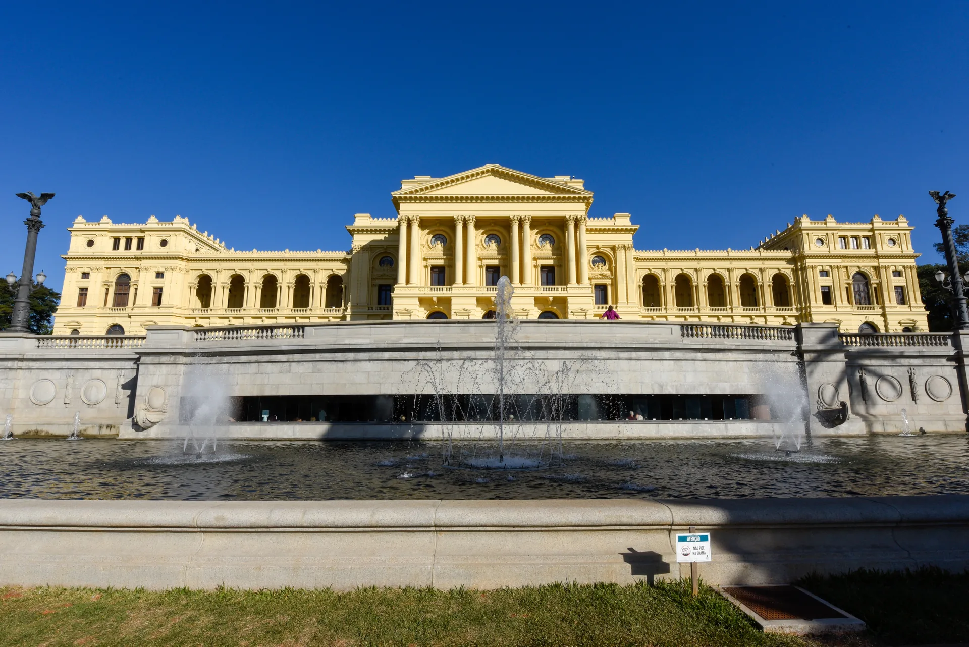 Museu do Ipiranga visto de frente, acima do espelho d'água e das fontes, sob céu azul intenso