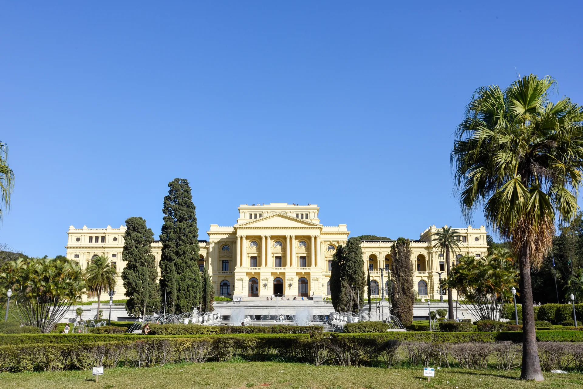 Fachada amarela do Museu do Ipiranga ao fundo dos jardins, entre ciprestes e uma palmeira, sob céu azul