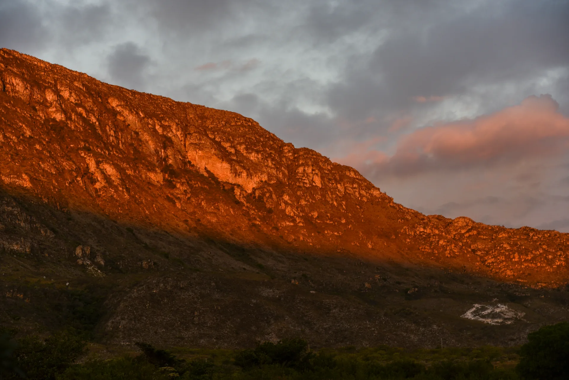 Paredão de pedra iluminado em laranja pelo sol do fim de tarde, sob nuvens cinzentas, em Lapinha da Serra