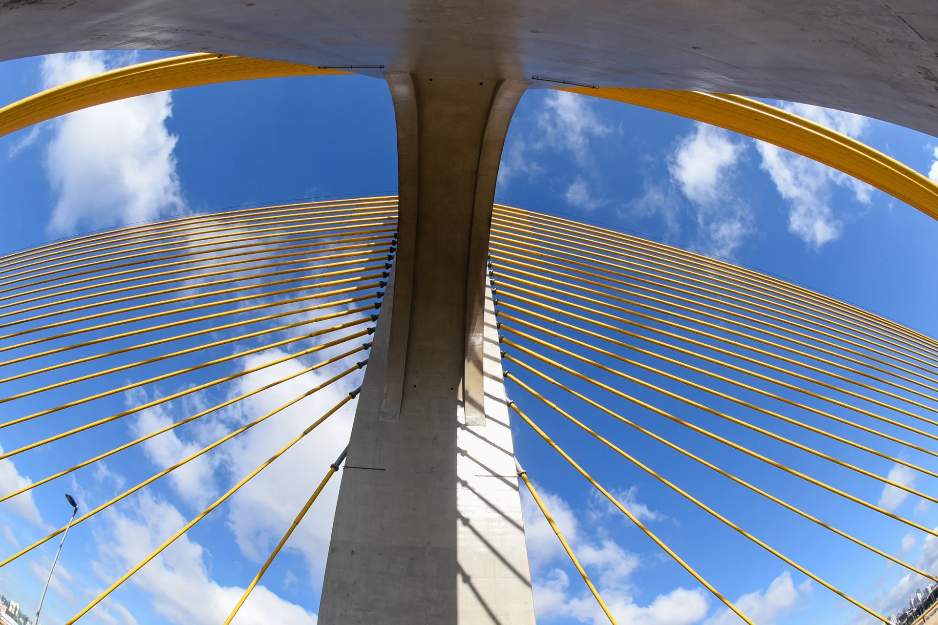Mastro de concreto e cabos amarelos de uma ponte estaiada vistos de baixo, contra céu azul com nuvens