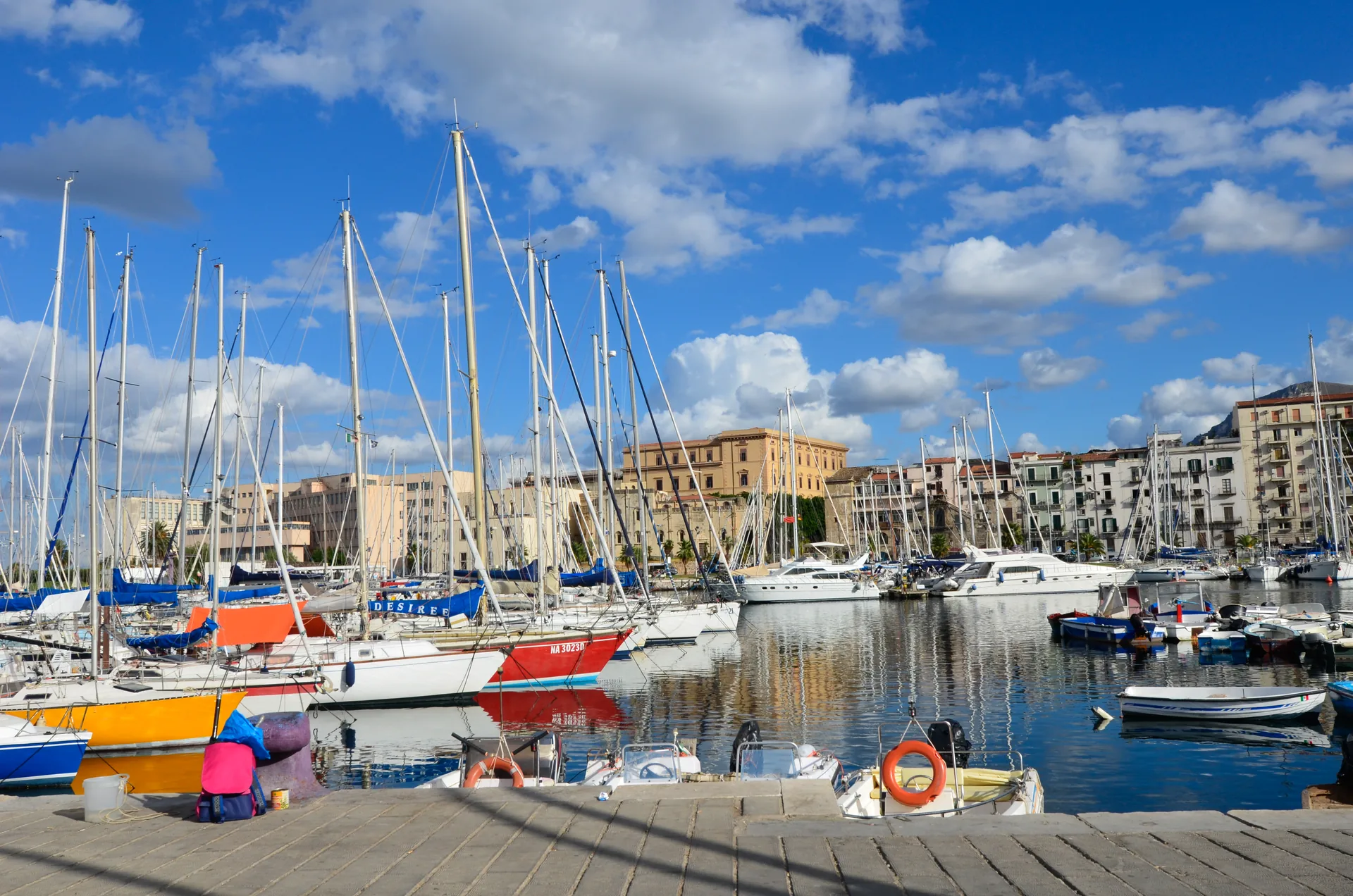 Veleiros e barcos ancorados no Porto de La Cala, com prédios antigos de Palermo ao fundo, sob céu azul
