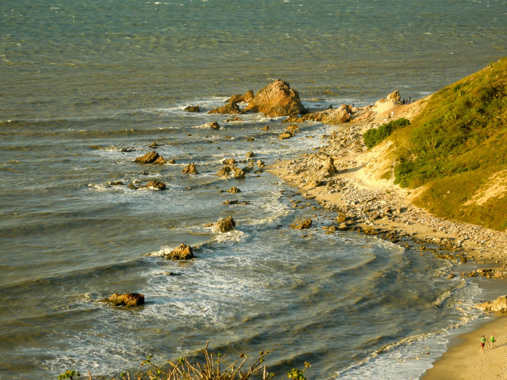 Mar com pedras e ondas junto a uma encosta verde, visto do alto do Morro do Serrote, em Jericoacoara